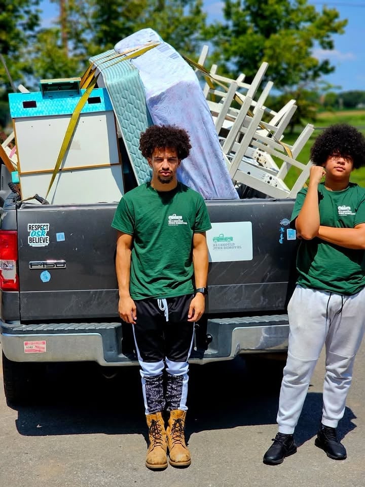 Two people in green volunteer shirts stand in front of a pickup truck loaded with household items and furniture for delivery