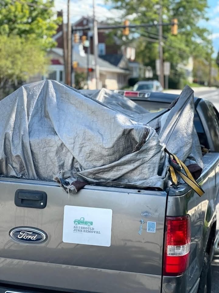 Gray Ford pickup truck with tarp covering bed and AIS Junk Removal decal on tailgate, parked on street with trees and buildings visible